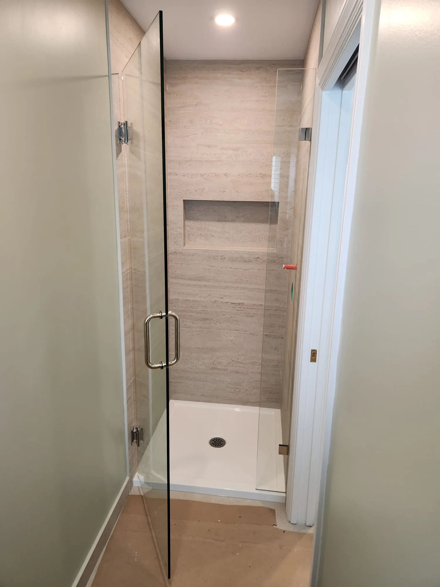 Frameless shower door ajar — showing door panel, chrome handle, and travertine tile surround