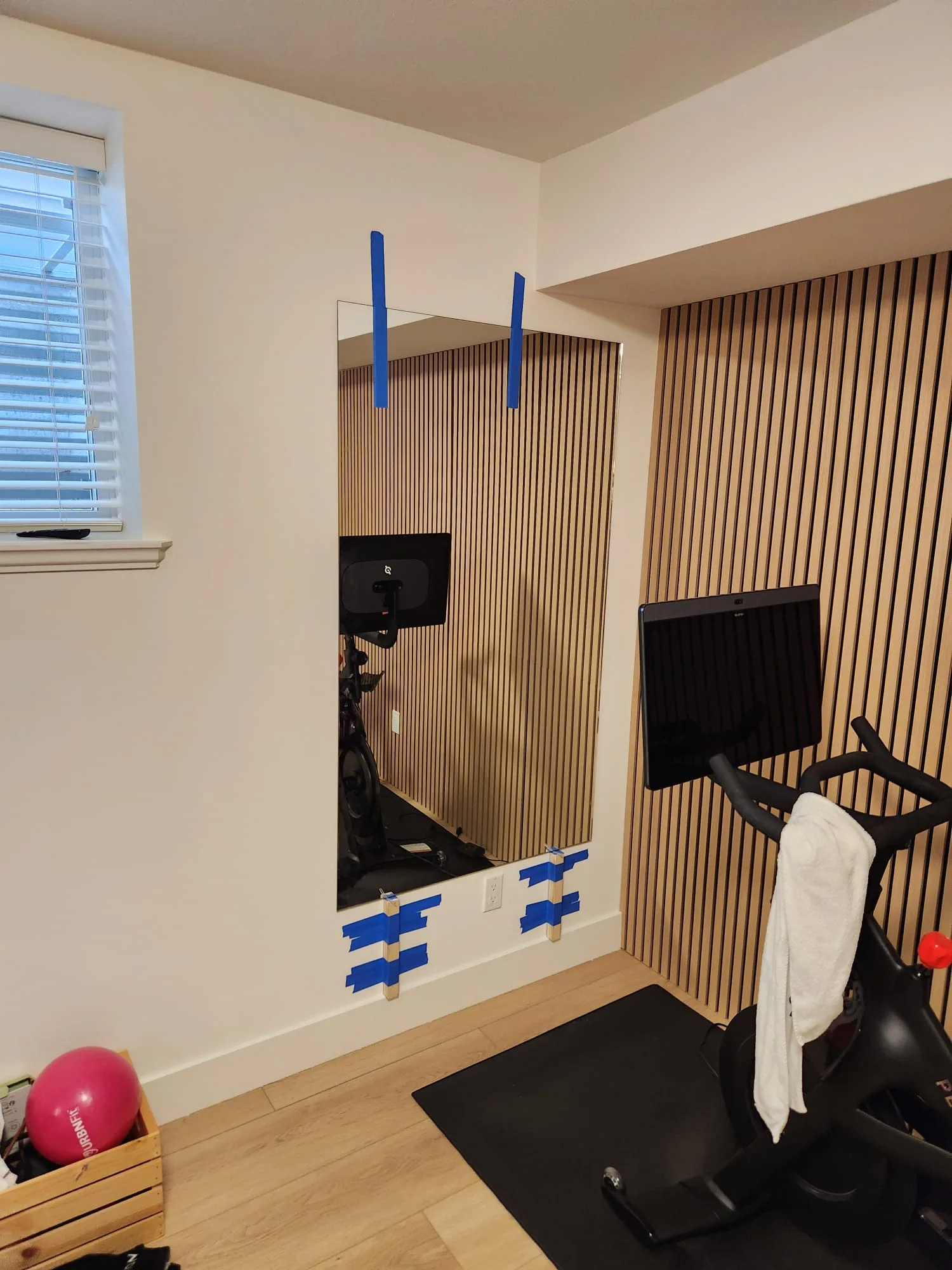 Mirror panel installation in progress — blue painter's tape holding panel while mastic cures, Peloton bike and slat wall accent visible
