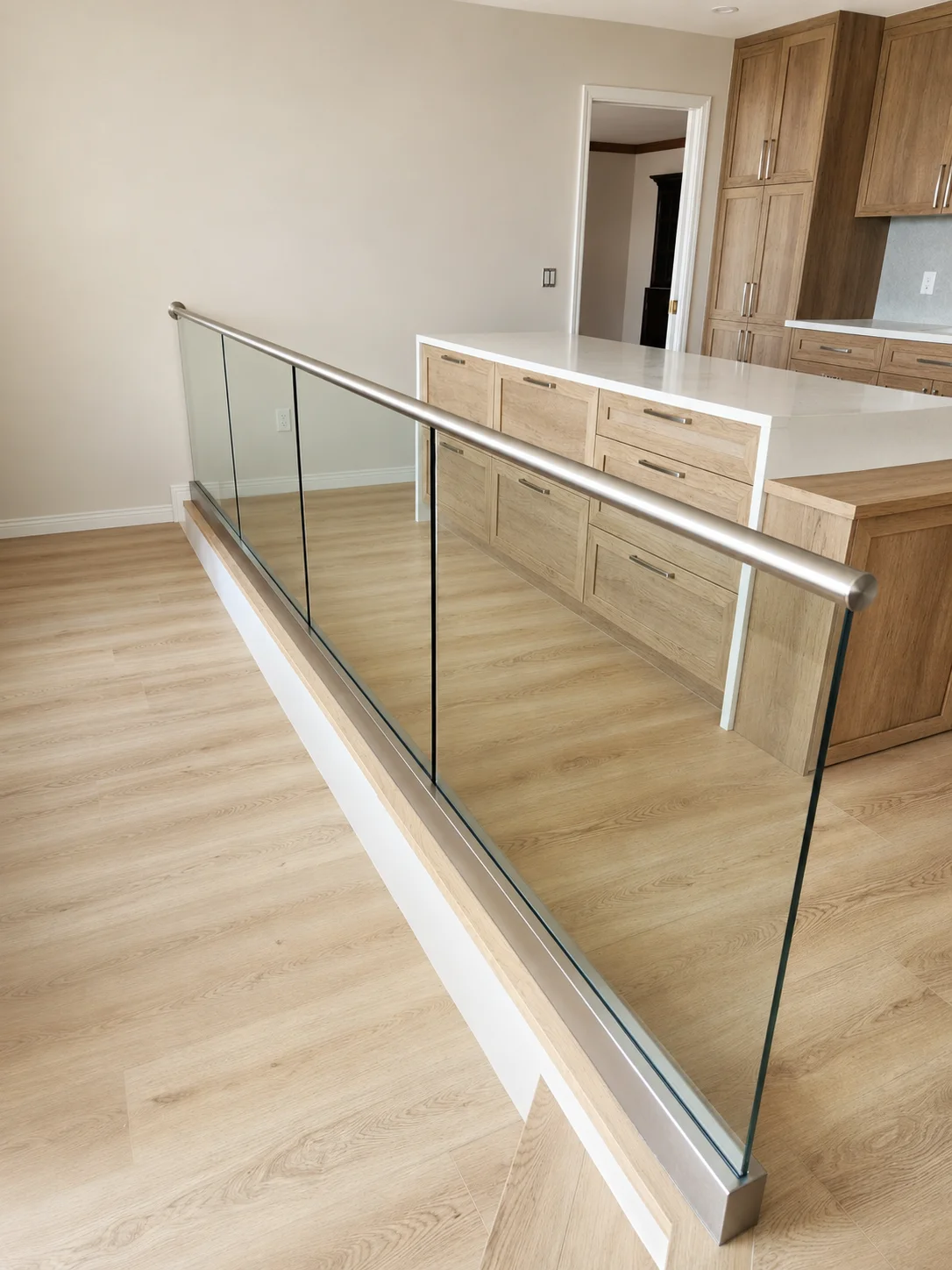 Close-up of base-shoe glass railing with brushed stainless round handrail — kitchen and living area visible beyond