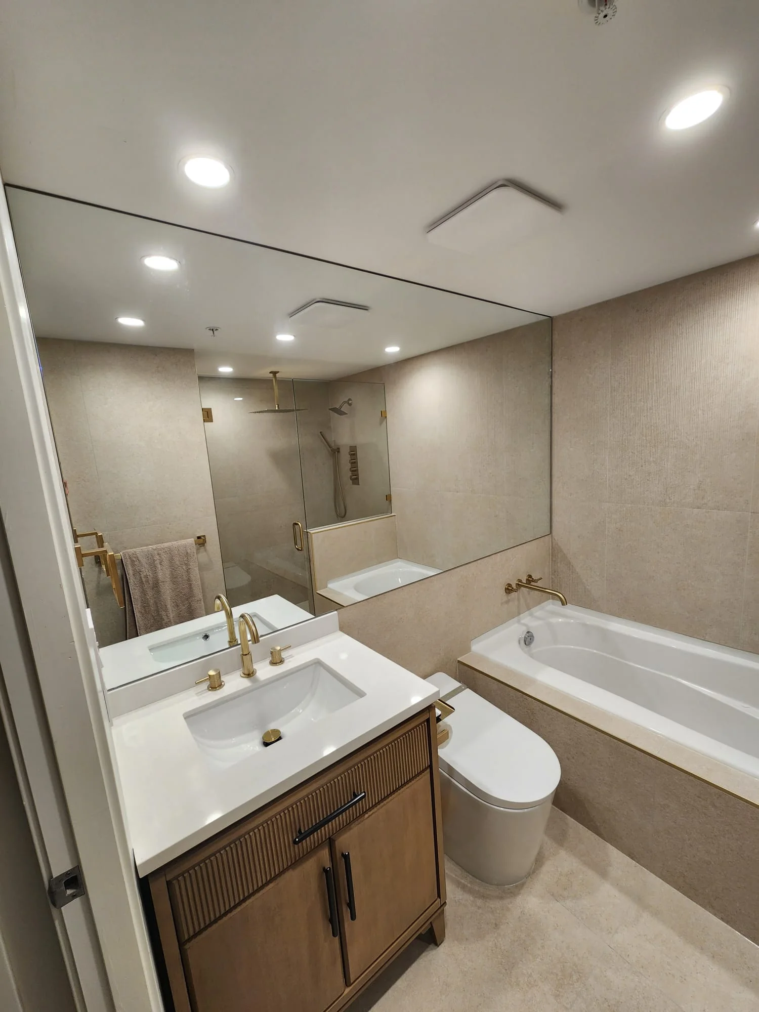 Full bathroom view — large full-width frameless polished mirror spanning vanity wall, brushed gold faucet, fluted wood vanity cabinet, soaker tub with wall-mount gold faucet