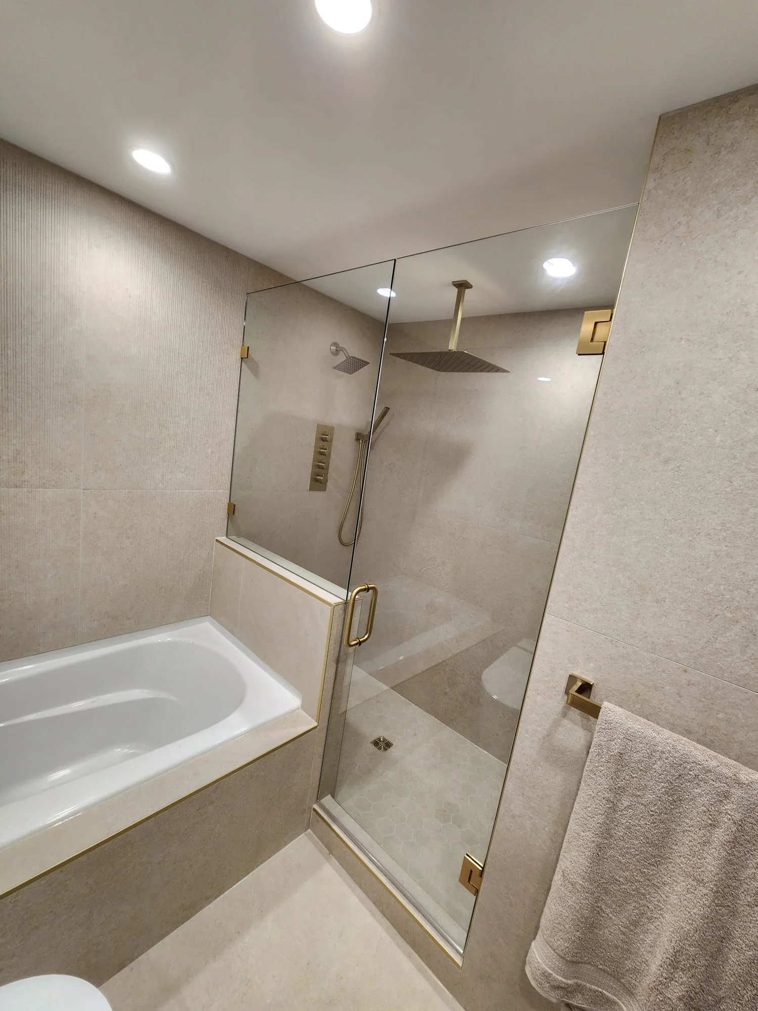 Full-height view of frameless shower enclosure from the soaker tub side — gold hardware, ceiling rain head, hexagonal tile floor drain visible
